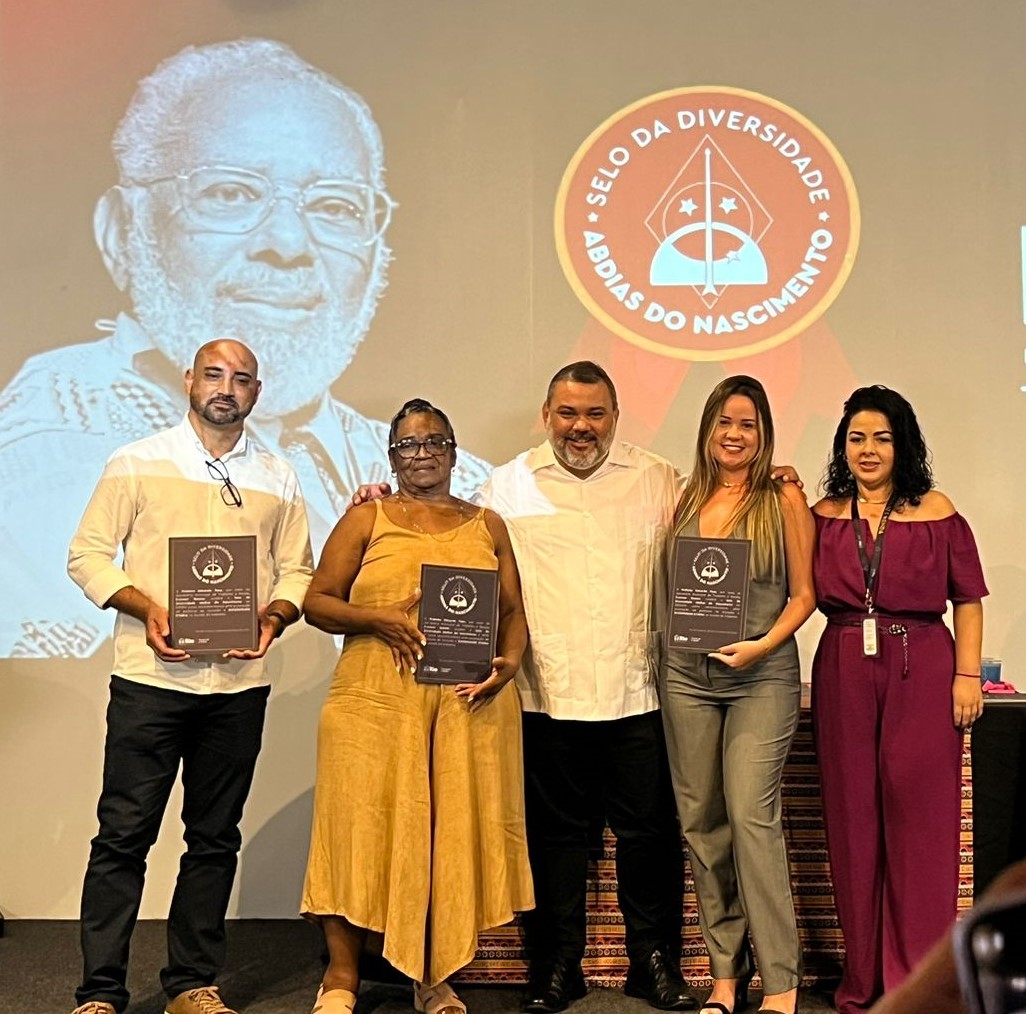 Fotografia da equipe premiada durante a cerimônia do Selo da Diversidade Abdias do Nascimento. Representantes seguram suas placas de reconhecimento em frente a um banner do evento, com a imagem de Abdias do Nascimento ao fundo. O momento celebra o impacto positivo das iniciativas inclusivas promovidas pelas instituições homenageadas.